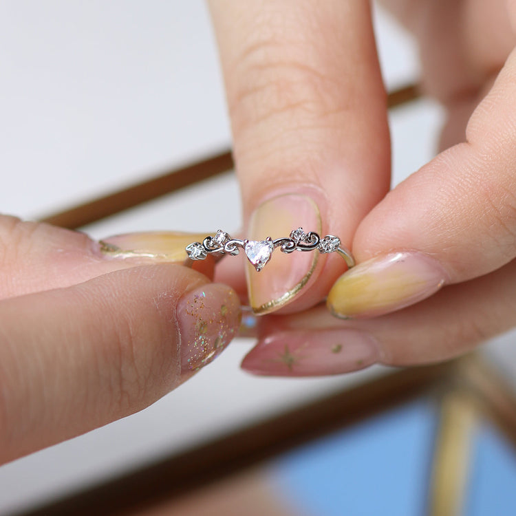 Close-up of an elegant heart ring held between fingers, showcasing its delicate design and shimmering stones.